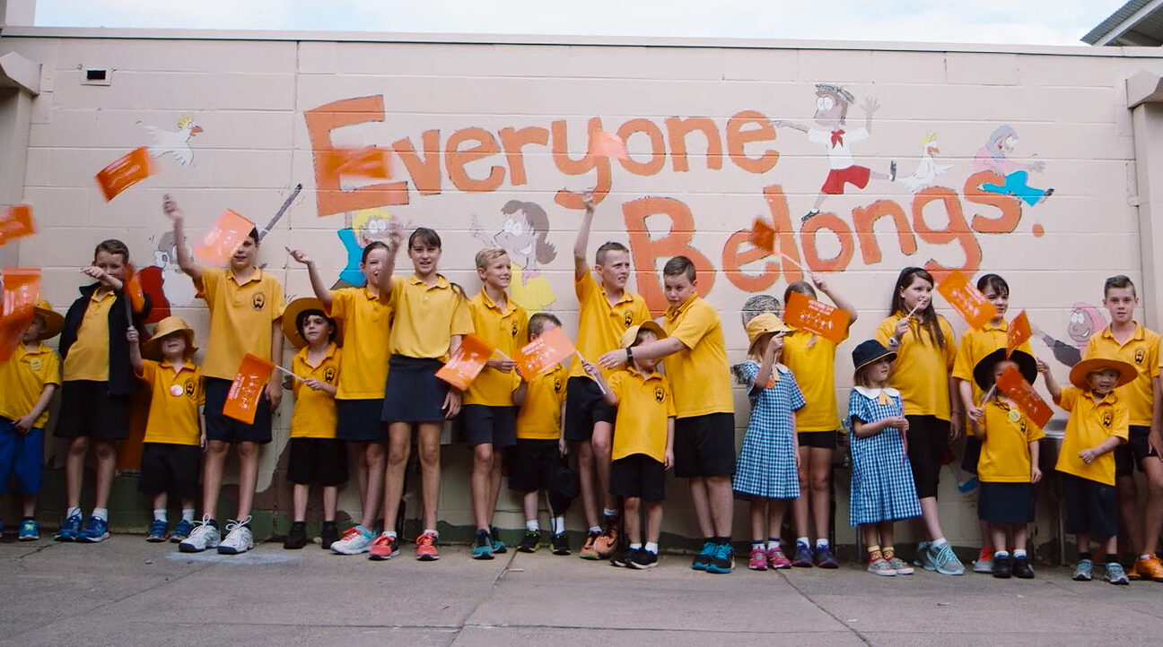 School children waving harmony day flags in front of wall with writing: Everyone belongs